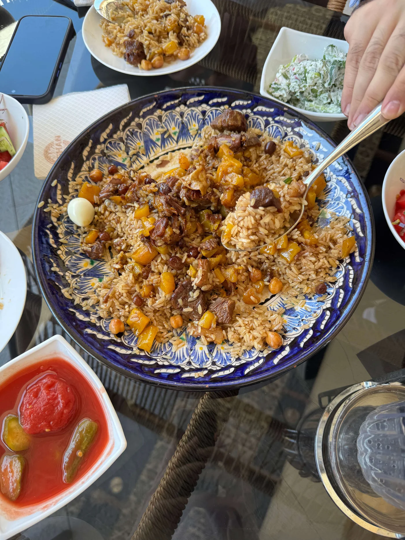 A large decorative bowl is filled with rice pilaf mixed with diced vegetables and pieces of meat. A hand holds a spoon, serving from the dish at a glass-topped table. Small side dishes with pickled vegetables, salad, and yogurt-based dip surround the main plate. The scene suggests a casual shared meal in a bright, relaxed setting.