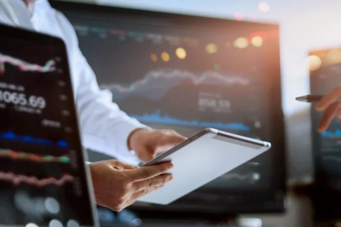 Personen nutzen ein Tablet, um Aktienmarktdiagramme und Finanzdaten zu analysieren, während im Hintergrund große Monitore mit Grafiken zu sehen sind.