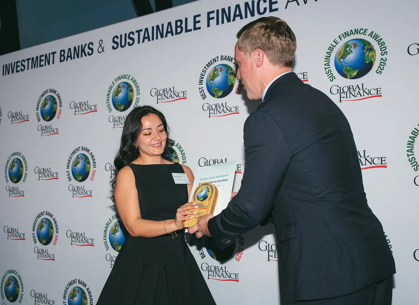 Eine Person in festlicher Kleidung erhält eine Trophäe und eine Urkunde bei einer Sustainable Finance Awards Veranstaltung mit gebrandeter Fotowand.