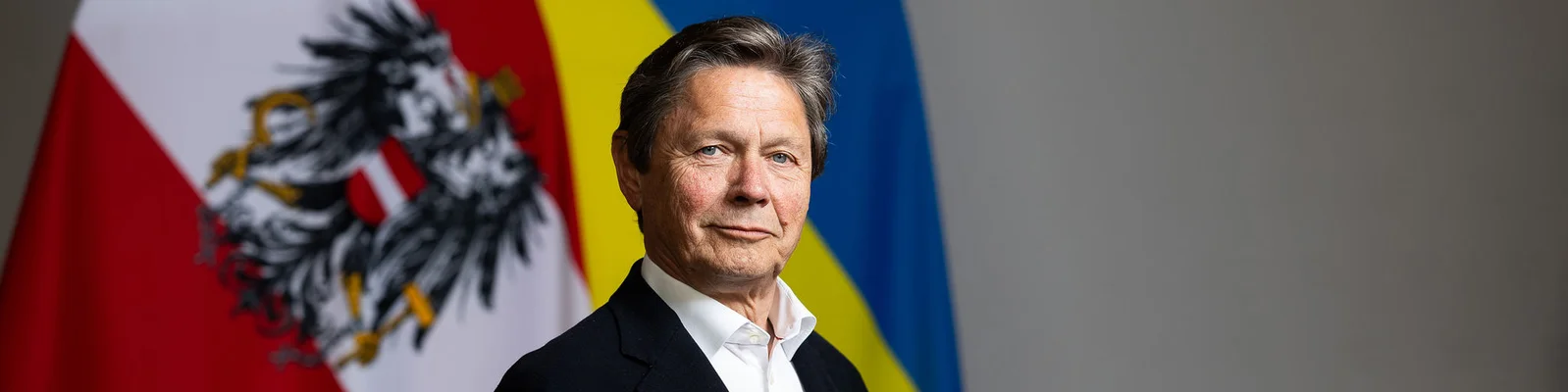 Eine Person in formeller Kleidung steht vor zwei großen Flaggen, einer österreichischen und einer ukrainischen. Die Szene wirkt offiziell oder diplomatisch, der Hintergrund ist neutral. Die österreichische Flagge zeigt einen markanten schwarzen Adler, die ukrainische ihre charakteristischen blauen und gelben Streifen. Die Stimmung ist insgesamt formell und ernst, der Fokus liegt auf den internationalen Beziehungen.