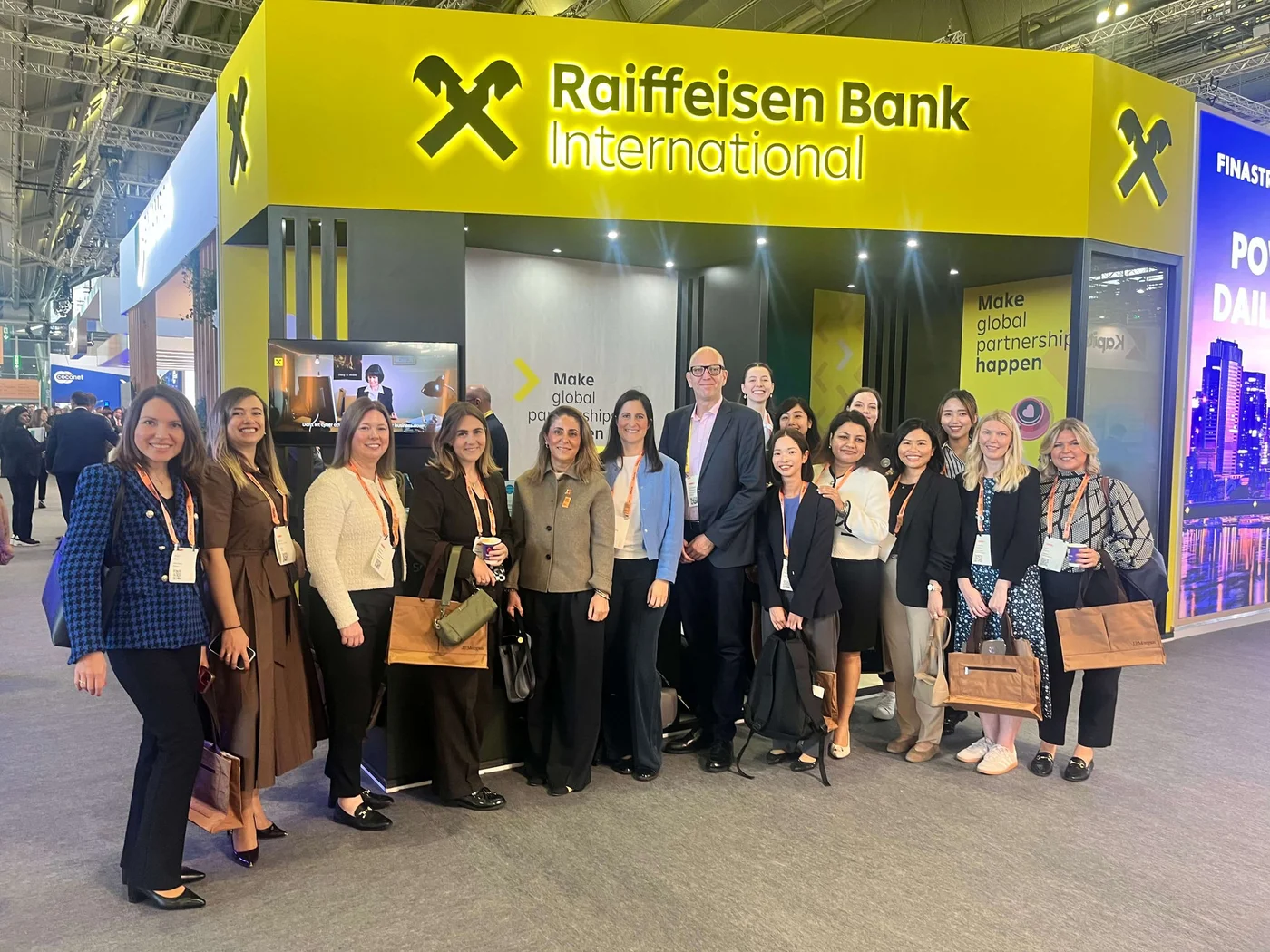 A group of professionals poses in front of the Raiffeisen Bank International booth at a corporate event. The booth features bright yellow branding and the slogan 'Make global partnerships happen.' The setting appears to be an indoor exhibition space with other booths visible in the background. The group is dressed in formal attire, holding conference bags.