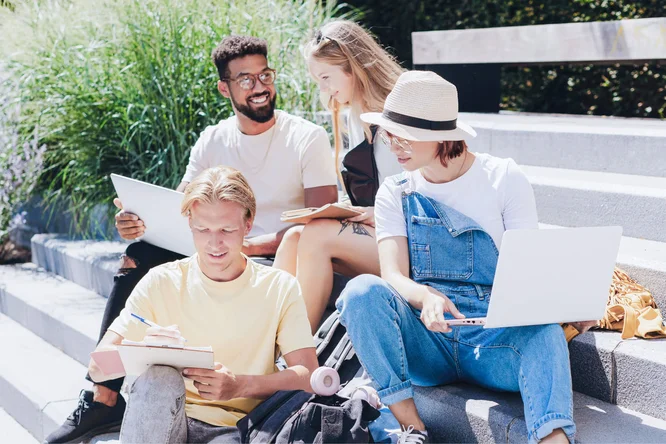 A group of students engaged in collaborative study outdoors, utilizing laptops and notebooks. The setting on stairs surrounded by greenery provides a relaxed and inspiring environment for learning and discussion.