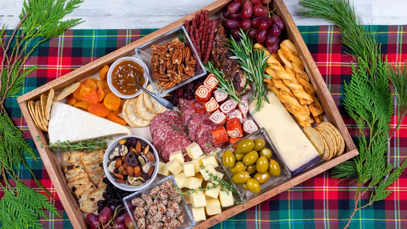 Eine große Holzplatte mit Käse, Wurst, Nüssen, Trockenfrüchten, Oliven und Crackern, dekoriert mit Grünzeug auf einem bunten Karo-Tuch.