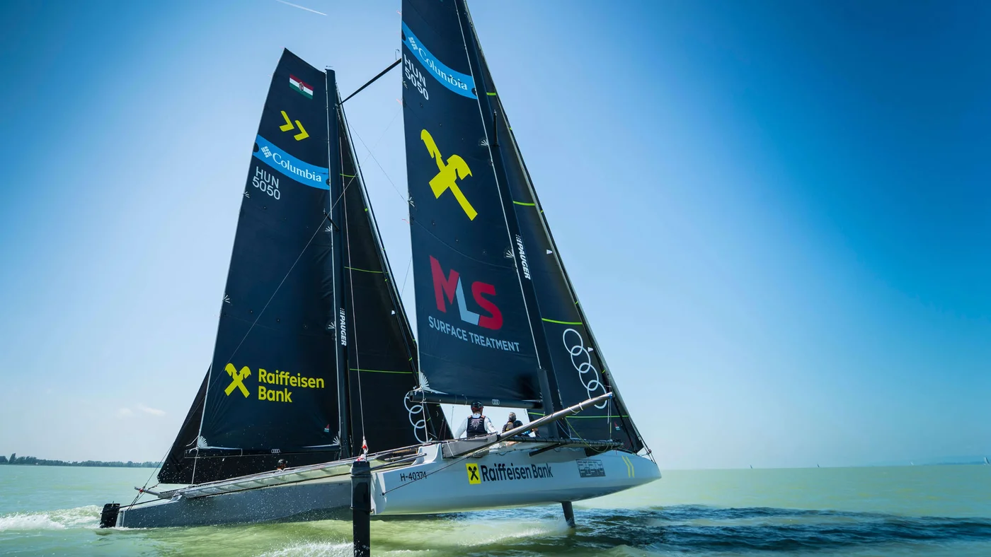 A sleek sailing catamaran glides across calm waters under a bright blue sky. The sails prominently display logos and text, including 'Raiffeisen Bank' and 'MS Service Treatment.' The scene captures the dynamic motion of the boat and the vibrant colors of the branding. The setting suggests a professional or competitive sailing activity.