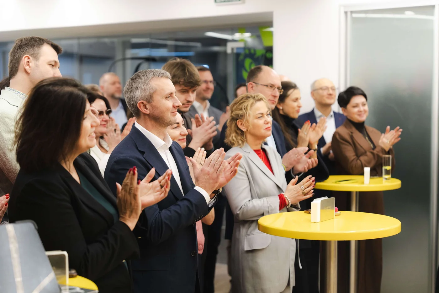Eine Gruppe Erwachsener in Businesskleidung steht in einem modernen Innenraum um gelbe Tische und applaudiert. Die Atmosphäre lässt auf ein Firmen- oder Netzwerk-Event schließen, mit heller Beleuchtung und zeitgemäßer Einrichtung. Die Stimmung ist positiv und engagiert; die Teilnehmer zeigen Wertschätzung und Unterstützung. Das Bild enthält keine sichtbaren Texte oder Zahlen.
