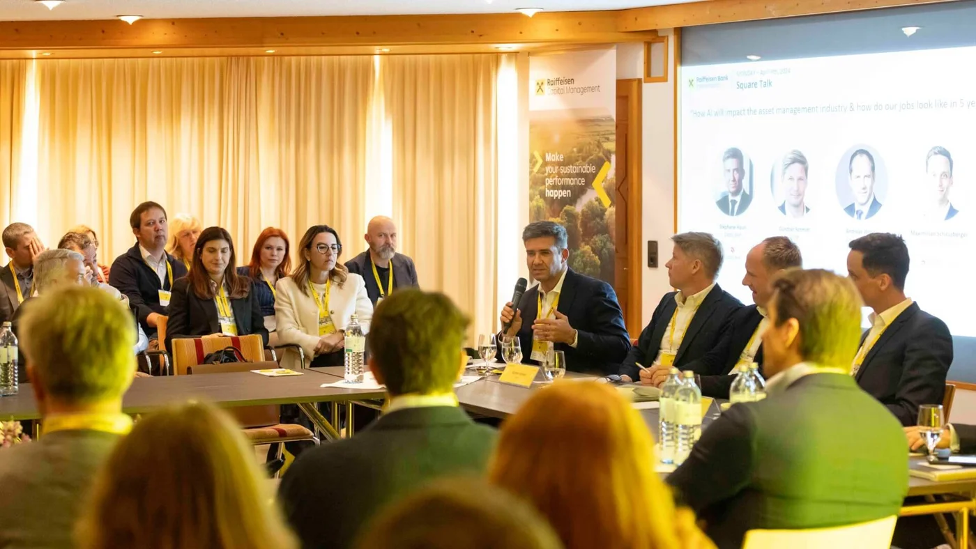 Eine Gruppe von Fachleuten sitzt an einem Tisch, wobei eine Person in ein Mikrofon spricht, während einer formellen Konferenz oder Podiumsdiskussion. Der Hintergrund zeigt einen Bildschirm mit Präsentationsfolien und Branding der Raiffeisen Bank.