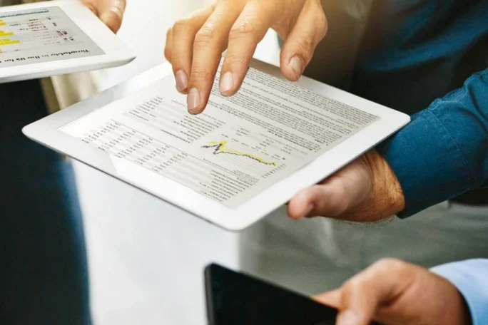 Hände halten und bedienen ein Tablet mit Finanzdiagrammen und Daten während eines Geschäftstreffens.