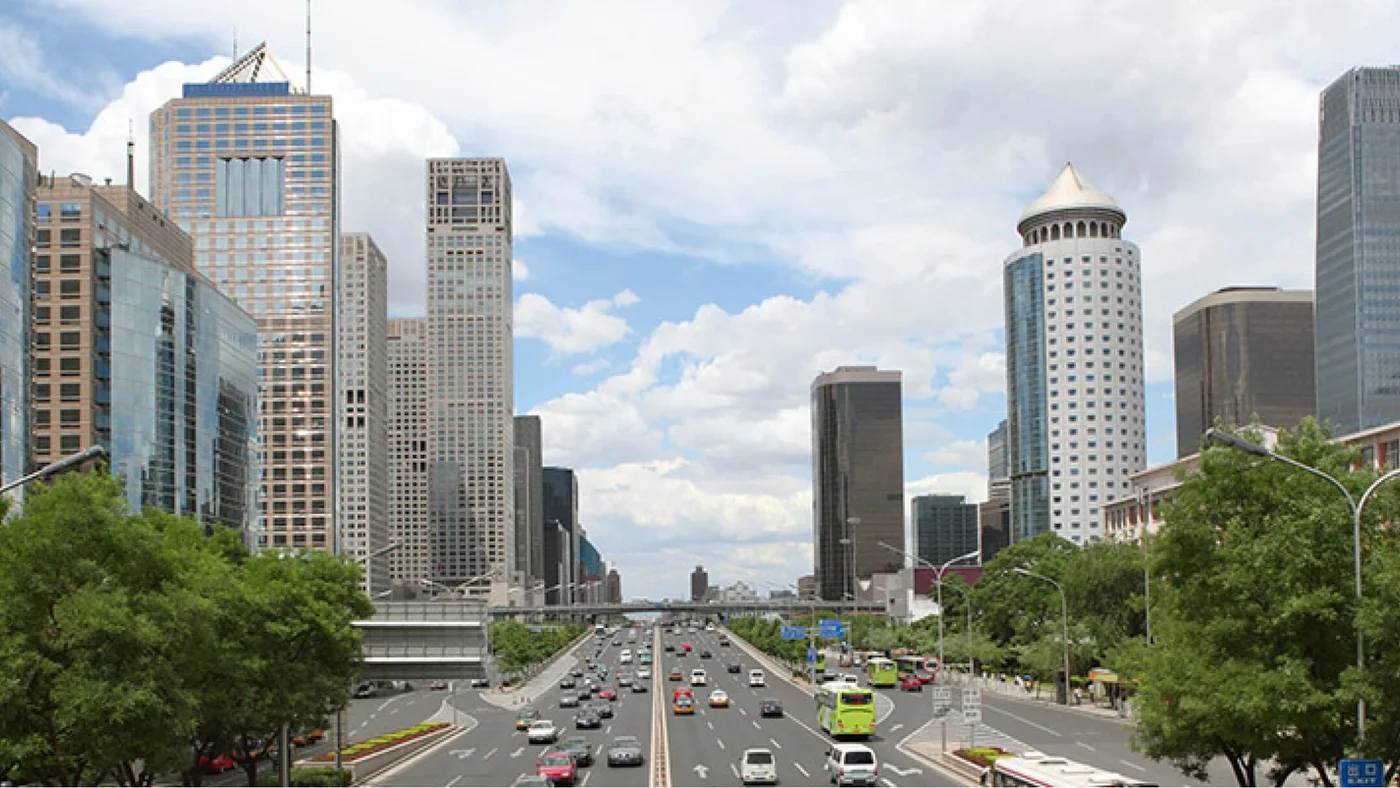 Lebendige urbane Szene mit einer belebten Straße, gesäumt von modernen Hochhäusern, fahrenden Fahrzeugen und üppigem Grün an den Seiten unter einem teilweise bewölkten Himmel.