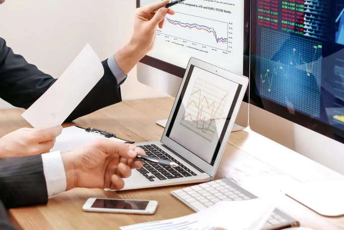 Fachleute führen eine ausführliche Diskussion und analysieren Finanzdaten und Markttrends, die auf mehreren Monitoren angezeigt werden, wobei der Schwerpunkt auf strategischer Planung und Entscheidungsfindung liegt.