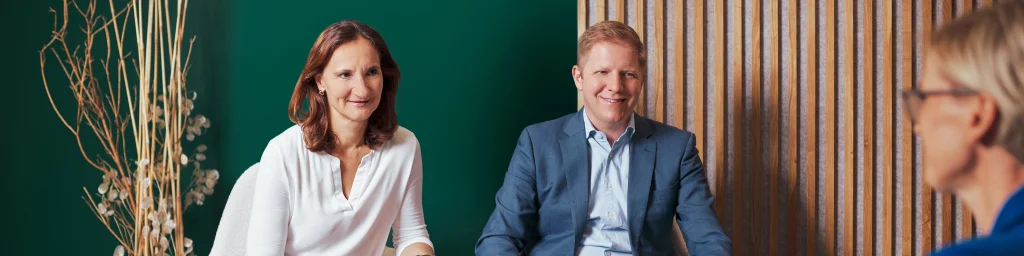 Professionelle Besprechungsszene mit drei Personen, die in einem modernen Büro sitzen. Im Hintergrund befinden sich eine grüne Wand und Holzvertäfelung, ergänzt durch minimalistische Dekoration mit Trockenpflanzen. Die Personen sind formell gekleidet, was auf ein unternehmerisches oder geschäftliches Umfeld hindeutet.