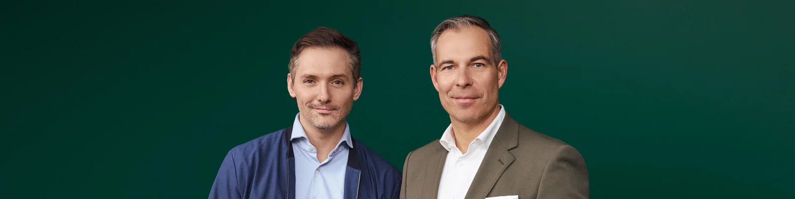 Henry Murray von Waterdrop und Daniel Rath sind formell gekleidet: Er trägt einen blauen Blazer und ein blaues Hemd, er einen braunen Anzug mit weißem Hemd. Der einfarbig grüne Hintergrund erzeugt eine professionelle und klare Bildwirkung. Das Foto unterstreicht den geschäftlichen oder formellen Charakter des Bildes.