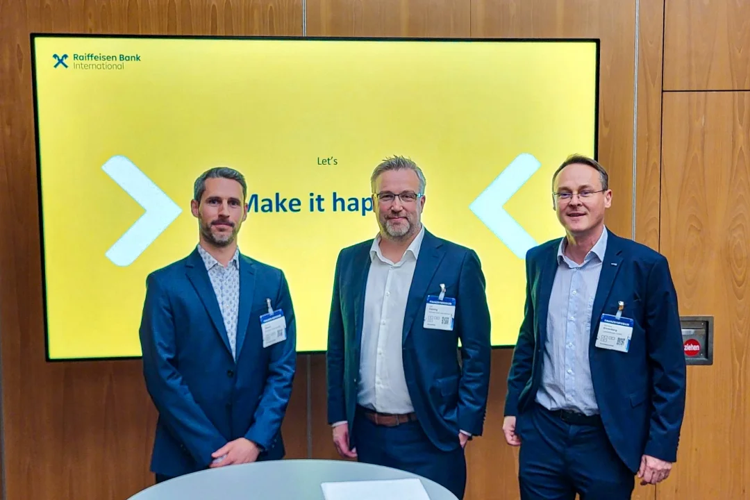 Experten der Raiffeisen Bank International stehen vor einem Präsentationsbildschirm während einer Konferenz, auf dem der Slogan "Make it happen" angezeigt wird