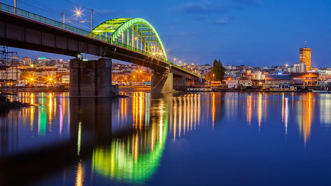 Eine wunderschön beleuchtete Brücke in Belgrad spiegelt lebendige Lichter im ruhigen Fluss darunter wider, mit der Stadtsilhouette im Hintergrund in der Dämmerung.