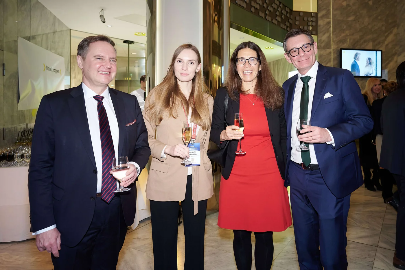 Vier elegant gekleidete Personen stehen gemeinsam bei einem Firmenevent und halten jeweils ein Glas Wein oder Sekt.