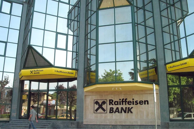 Der Eingang der Raiffeisen Bank Albania mit großen Glasfenstern und gelber Beschilderung. Das moderne architektonische Design spiegelt die Umgebung wider.