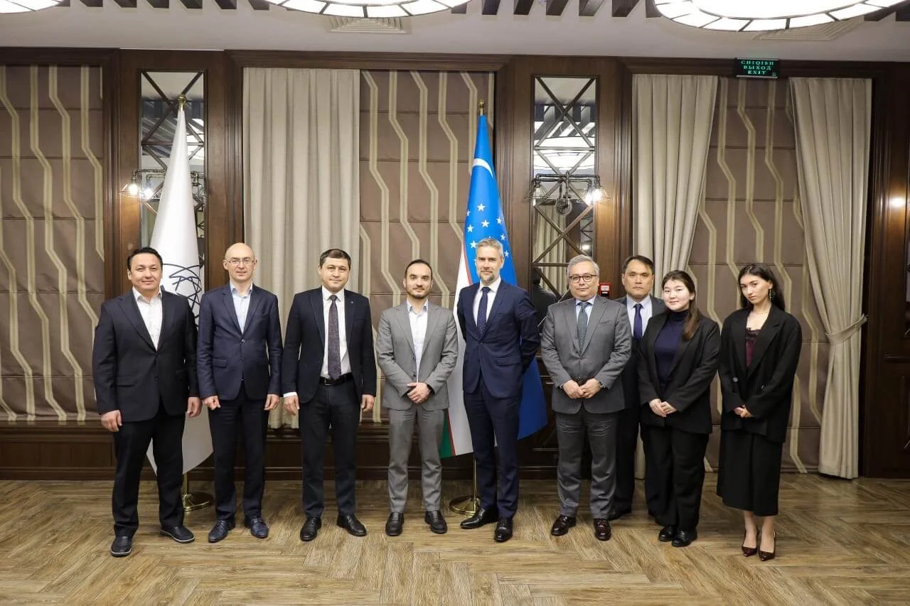 A group of professionally dressed adults stands in a line for a formal group portrait. The setting appears to be an indoor conference or meeting room with patterned curtains and wooden paneling. A blue national flag with yellow stars and white stripes is prominently displayed behind the group. The individuals wear business suits and smart attire, suggesting a diplomatic or corporate event.