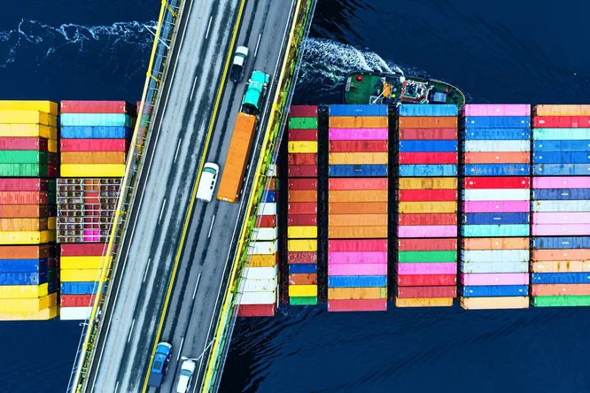 Eine Draufsicht auf eine Brücke mit Autos, die über ein großes Frachtschiff fahren, das bunte Schiffscontainer auf dunkelblauem Wasser transportiert.