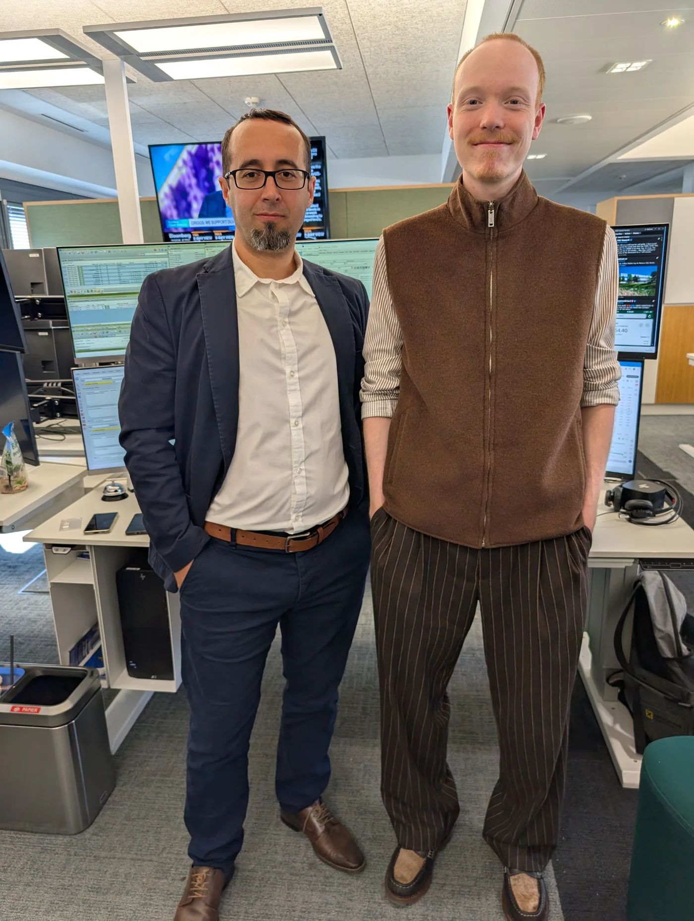 Two colleagues stand side by side in a contemporary office environment. One wears a navy suit with a white shirt, while the other is dressed in a brown sleeveless sweater and striped trousers. Behind them are multiple computer monitors displaying data and a wall-mounted television screen. The workspace features desks, office chairs, and various electronic equipment, suggesting a professional, technology-driven setting.
