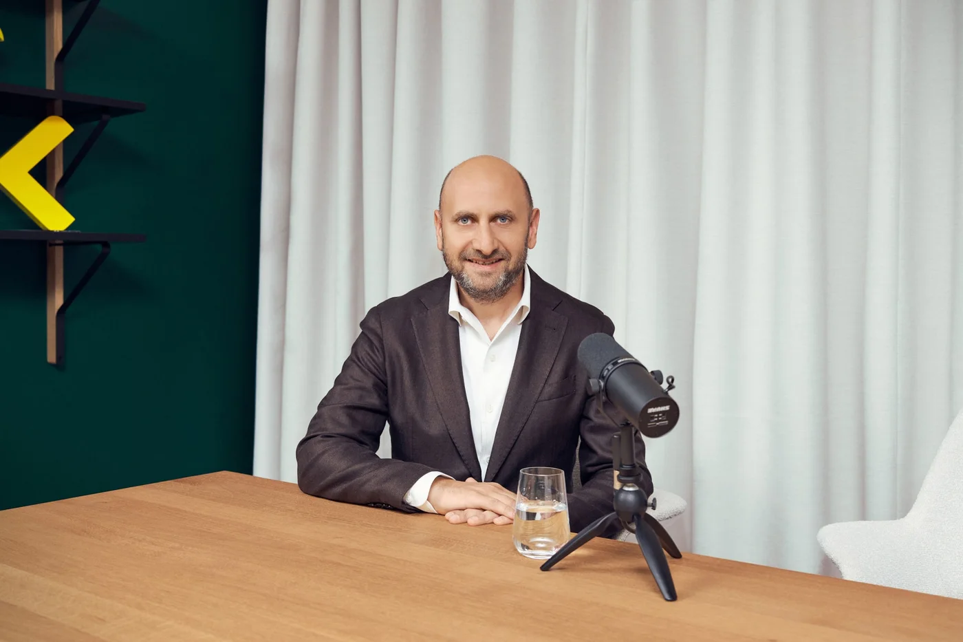 A bald male professional in a dark suit sits at a wooden table in front of a microphone, ready to speak. The setting appears to be a modern studio or office with a green wall and white curtains in the background. A glass of water is placed on the table near his hands, and a white chair is partially visible to the side. The overall scene suggests a podcast, interview, or recorded conversation in a clean, contemporary environment.