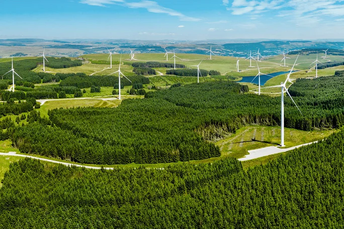 Luftaufnahme eines Windparks mit mehreren Windkraftanlagen vor einer üppig grünen Landschaft und klarem, blauem Himmel.