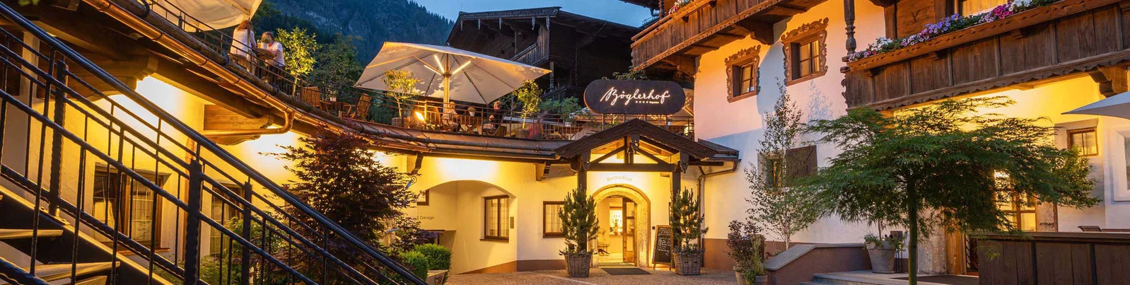 An inviting courtyard of an alpine-style building illuminated by warm lights during dusk. The setting features wooden architecture, outdoor seating with umbrellas, and lush greenery. A visible sign reads 'Alpenhotel,' adding to the charm of the scene.