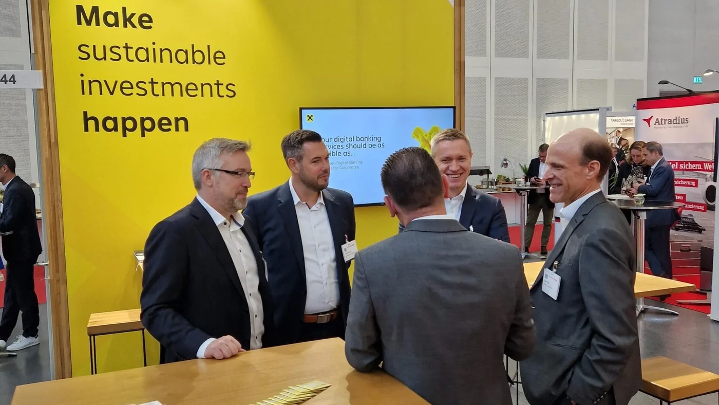 An einem Messestand mit einer gelben Wand, auf der steht: „Machen Sie nachhaltige Investitionen möglich“, unterhalten sich mehrere Personen in Businesskleidung.