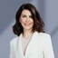 Portrait of Yulia Shults smiling confidently in a light-colored textured blazer over a neutral top. She has shoulder-length dark brown hair styled in soft waves. The background is a clean, blurred gradient in cool gray tones, giving a modern and professional feel. The overall composition suggests a corporate or business profile image.