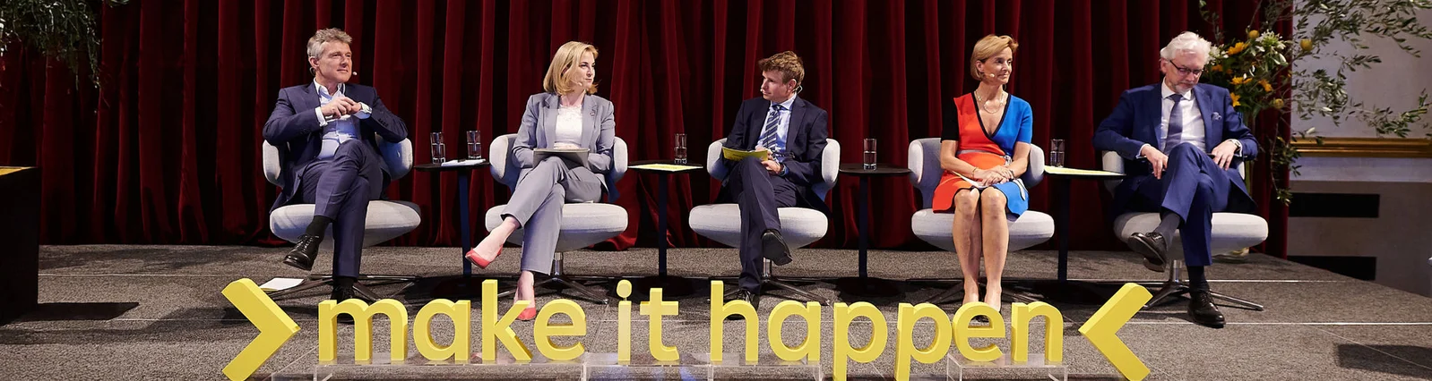 Eine Gruppe von fünf Personen sitzt während einer formellen Podiumsdiskussion auf der Bühne. Die Kulisse ist mit einem roten Vorhang und modernen Stühlen gestaltet. Vorne prangt in Gelb der Schriftzug „Make it happen“. Unter den Teilnehmern sind drei Männer und zwei Frauen. Sie tragen professionelle Kleidung, was an eine Firmen- oder Geschäftsveranstaltung erinnert.
