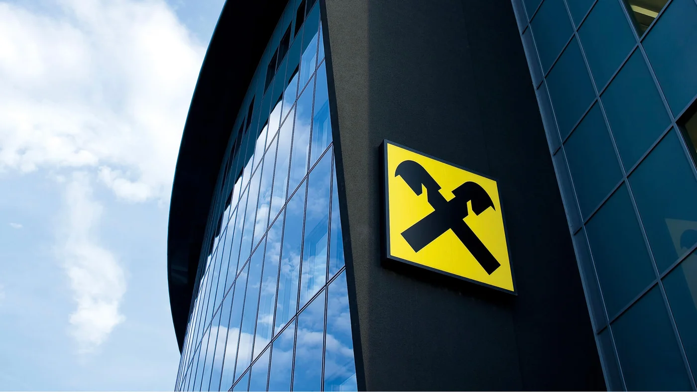 Moderne Gebäudefassade mit reflektierenden Glasfenstern und prominent platziertem Raiffeisen-Logo. Der Himmel spiegelt sich in den Fenstern wider und verleiht der Architektur eine dynamische Note.