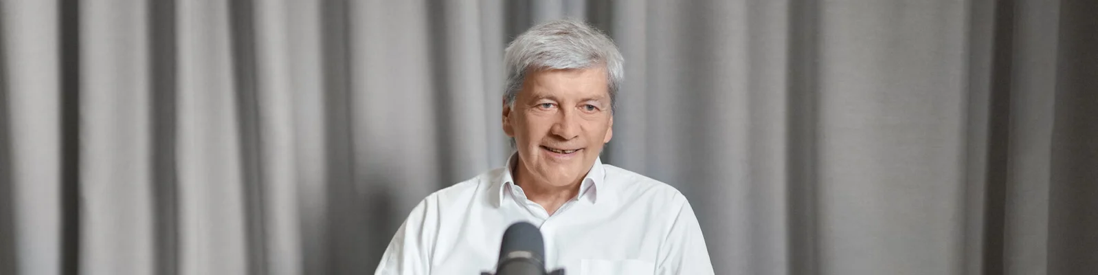 Dr. Johann Strobl trägt ein weißes Hemd und sitzt vor einem Mikrofon. Im Hintergrund sind graue Vorhänge zu sehen, was auf eine Innenumgebung wie ein Studio oder einen Interviewraum hinweist. Das Mikrofon ist deutlich sichtbar und deutet auf eine Aufnahme- oder Sprechsituation hin.