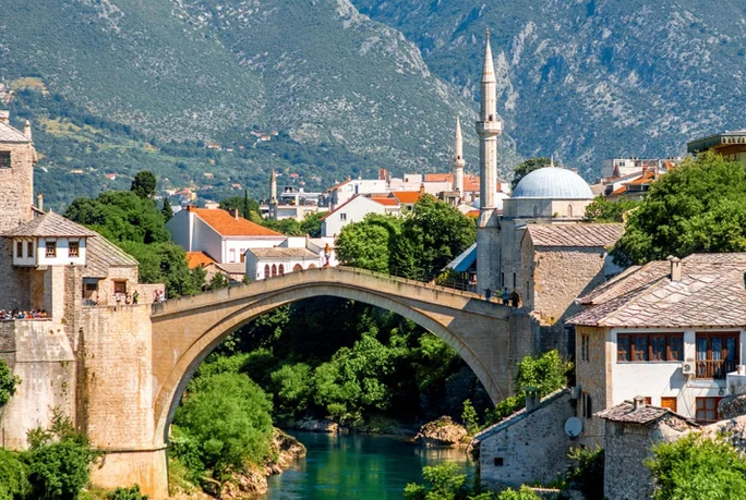 A picturesque view of a historic stone bridge spanning a turquoise river in a charming town surrounded by lush greenery and mountains. The setting includes traditional architecture with domed rooftops and minarets, evoking a cultural and serene atmosphere. The bright daylight enhances the vibrant colors of the landscape and buildings.