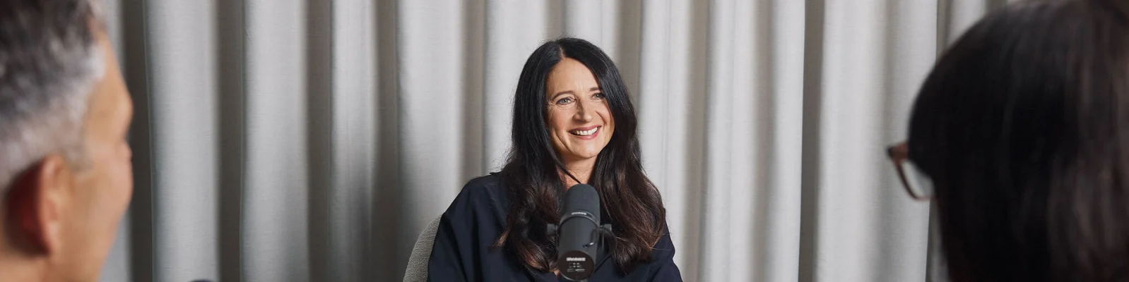 A podcast recording session featuring Marianne Heiß, a professional microphone setup in a studio environment. The setting includes neutral curtains in the background, creating a focused and intimate atmosphere. Two individuals are partially visible in the foreground, suggesting a conversational format.