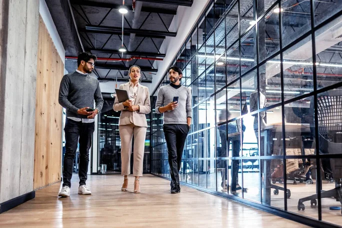 Drei Fachleute gehen durch einen stilvollen Büroflur mit Glaswänden, besprechen ihre Arbeit und verwenden digitale Geräte, was Teamarbeit, Innovation und ein modernes Geschäftsumfeld repräsentiert.