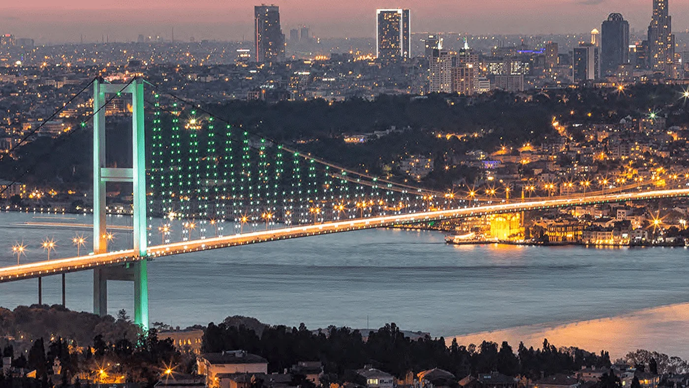 Atemberaubender Blick auf eine hell erleuchtete Brücke, die einen Fluss überspannt, mit funkelnden Stadtlichtern im Hintergrund während der Dämmerung.