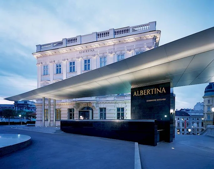 The Albertina Museum's modern entrance contrasts with its historic architecture in an evening setting. The illuminated facade highlights the building's classical design, while the sleek canopy adds a contemporary touch. The word 'Albertina' is prominently visible on the structure.