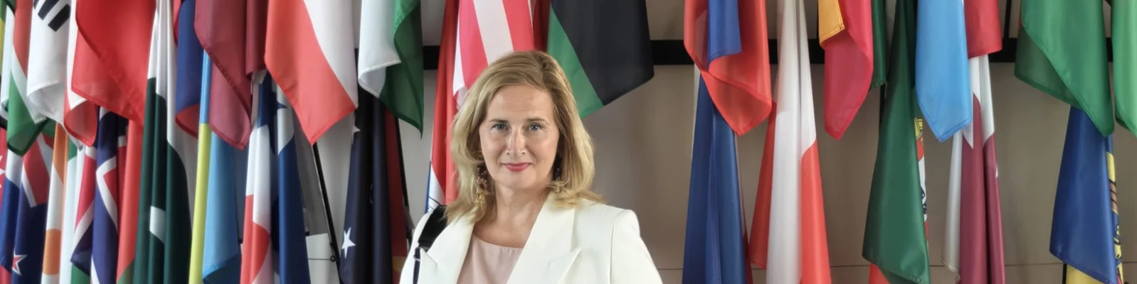 A professionally dressed woman stands in front of a display of numerous national flags. The setting appears to be an international conference or institutional lobby. Bright colors from the flags create a vibrant backdrop behind the subject. The overall scene suggests themes of diplomacy, global business, and international cooperation.