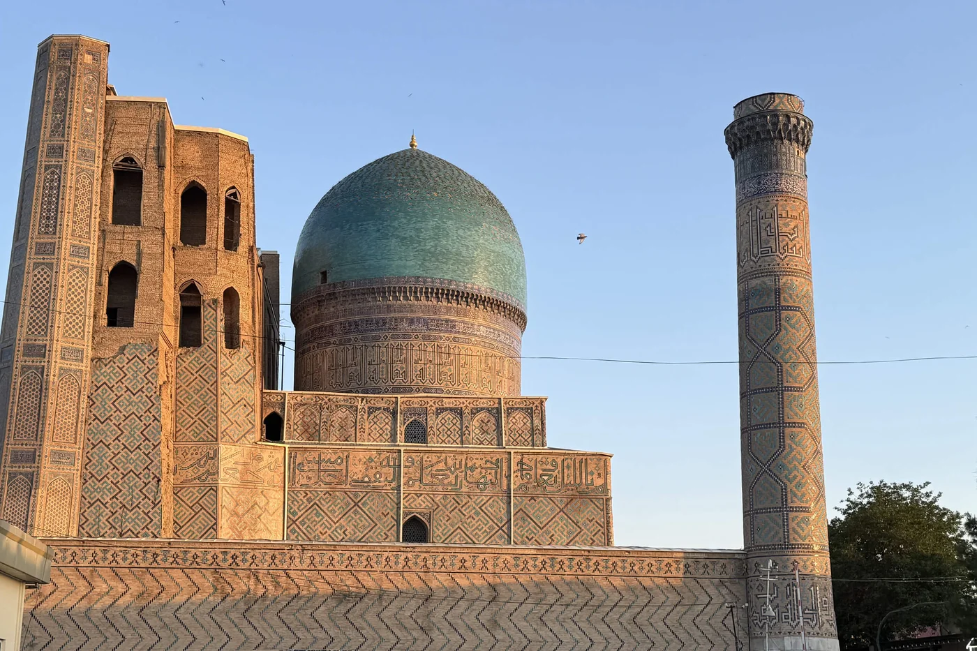 A historic mosque with a large turquoise dome rises against a clear evening sky. The building features intricate geometric tilework and decorative patterns in warm earth tones. A tall, slender minaret stands to the right, also covered in detailed ornamentation. Soft sunset light highlights the textures of the stone and tiles, while the surrounding area remains mostly in shadow.