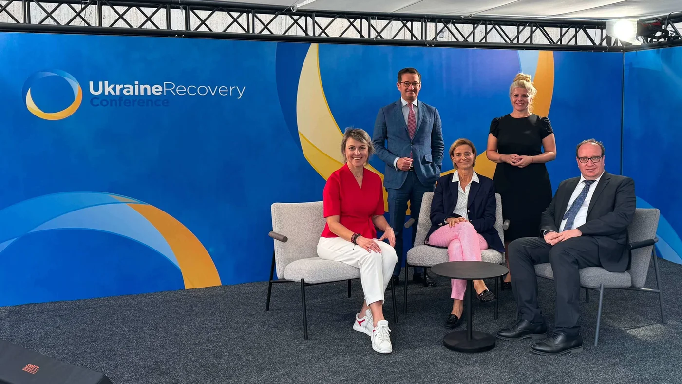 A group of five individuals is seated and standing in front of a blue and yellow backdrop with the text 'Ukraine Recovery Conference' clearly visible. The setting appears to be a formal event or conference. The individuals are dressed in business and semi-formal attire, and the stage setup includes chairs and a carpeted floor.