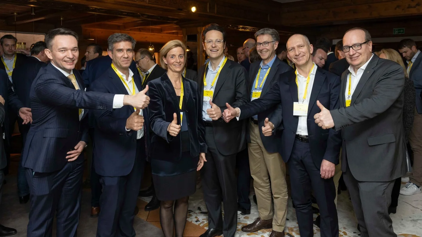 Eine Gruppe von Berufstätigen in Anzügen steht bei einer Konferenz zusammen, zeigt Daumen hoch und positive Gesten, mit vielen weiteren Personen im Hintergrund und Namensschildern um den Hals.