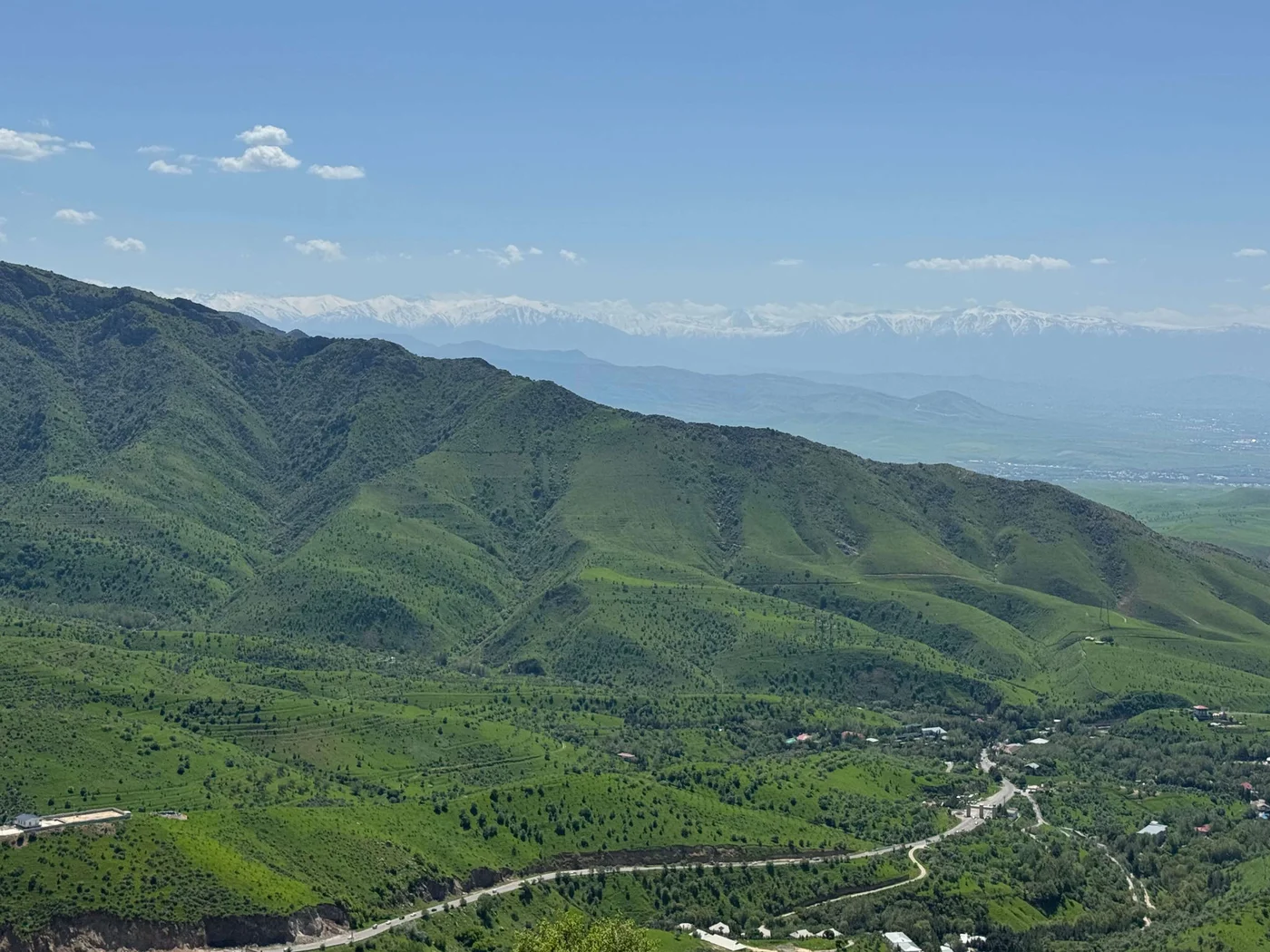 A vast green mountain range stretches across the scene under a bright blue sky. Rolling hills covered in dense vegetation dominate the foreground, with scattered houses and winding roads visible below. In the distance, hazy mountains form a soft backdrop, adding depth to the landscape. The overall atmosphere is peaceful and natural, highlighting the beauty of the countryside.