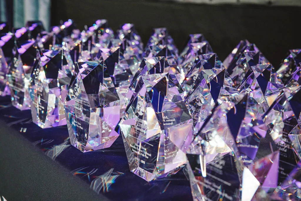 Multiple faceted crystal awards are arranged in neat rows on a dark tablecloth. The trophies catch the light, creating reflections and prismatic color highlights across their surfaces. Soft purple and blue lighting enhances the glassy texture and gives the scene a celebratory atmosphere. The background is slightly out of focus, keeping attention on the shimmering award shapes in the foreground.
