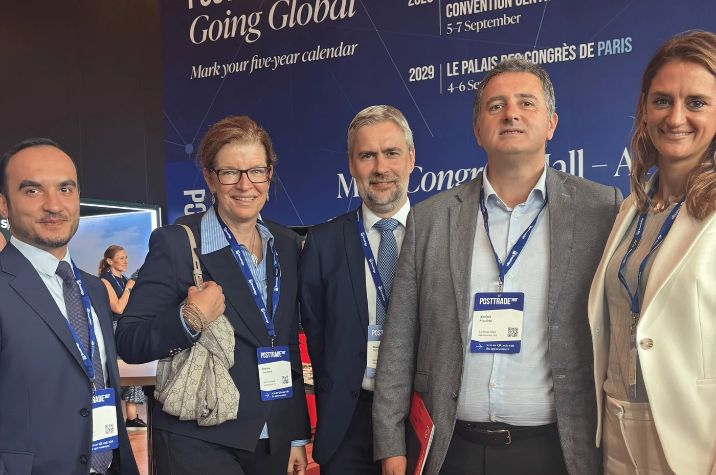 Eine Gruppe von fünf Geschäftsleuten posiert gemeinsam auf einer Konferenz. Sie tragen formelle Geschäftskleidung, Namensschilder und Lanyards sind gut sichtbar. Im Hintergrund ist eine große blaue Veranstaltungswand mit Texten zu sehen, die auf internationale Konferenzen und Termine, darunter das Jahr 2029, hinweisen. Die Szenerie vermittelt den Eindruck eines professionellen Netzwerkumfelds in einem modernen Kongresssaal.
