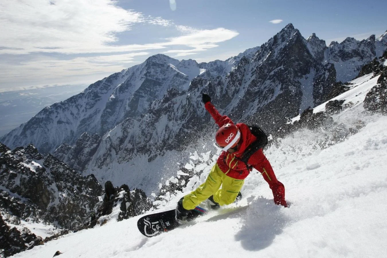 Ein Snowboarder in auffälliger Kleidung fährt eine steile, verschneite Piste in den Hohen Tatra mit schroffen Gipfeln im Hintergrund hinunter.