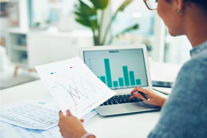Eine Person überprüft gedruckte Diagramme und Grafiken, während sie an einem Laptop mit Balkendiagrammen arbeitet. Die Umgebung ist eine moderne Büroumgebung mit Tageslicht und Grünflächen im Hintergrund. Der Fokus liegt auf Datenanalyse und beruflicher Produktivität.