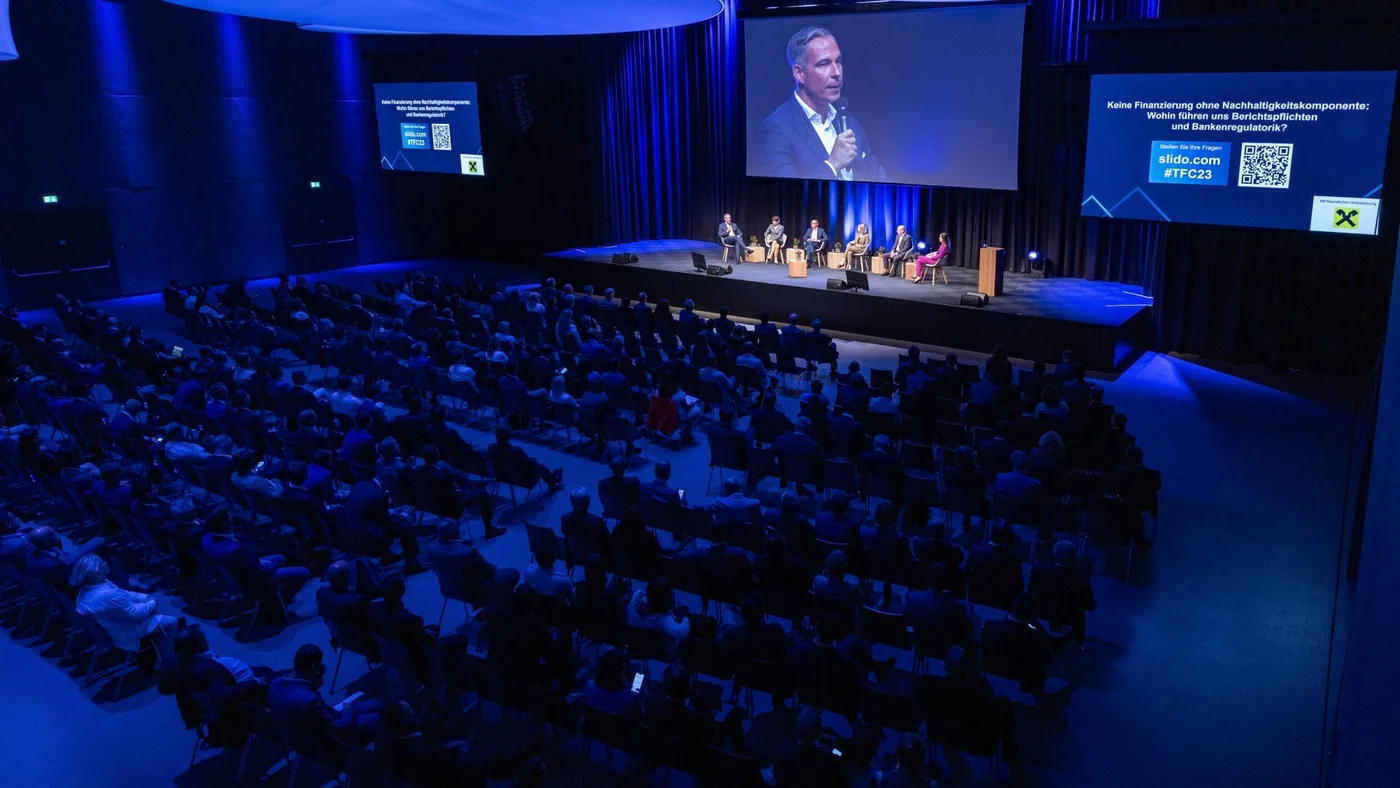 Eine Gruppe von Rednern sitzt auf der Bühne in einem großen Auditorium und wird vom Publikum beobachtet.