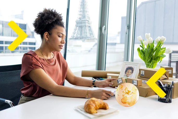 A woman with curly hair sitting at a desk in a bright office, with a view of the Eiffel Tower outside the window, surrounded by boxes, a framed photo, a vase of tulips, and a globe.