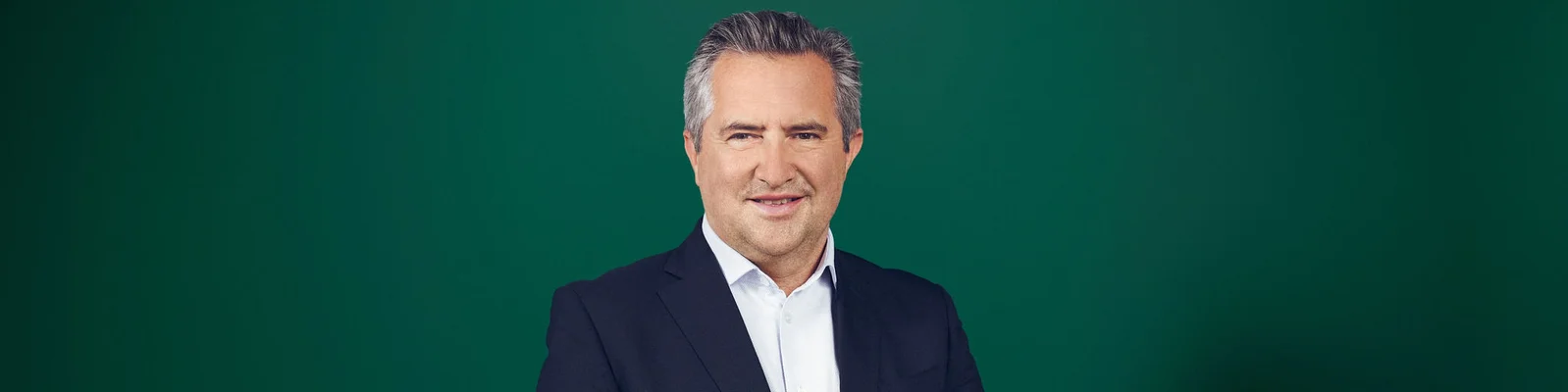 Rainer Schnabl, in dunklem Anzug und weißem Hemd, posiert vor einem einfarbig grünen Hintergrund. Mit einem leichten Lächeln blickt er in die Kamera und wirkt professionell und zugänglich. Das Bild ist klar und minimalistisch und konzentriert sich auf den Oberkörper und den Gesichtsausdruck des Porträtierten. Der schlichte Hintergrund unterstreicht den seriösen und formellen Charakter des Porträts.