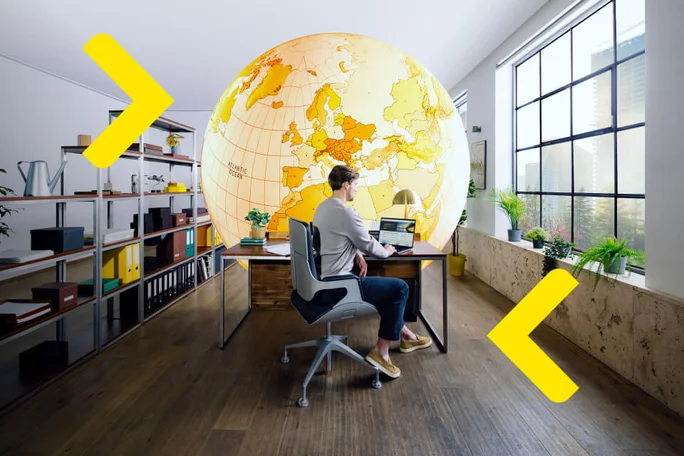 Eine Person sitzt an einem Schreibtisch und arbeitet an einem Laptop in einem hellen Büro, mit einer großen Weltkugel Europas und des Atlantiks an der Wand im Hintergrund.