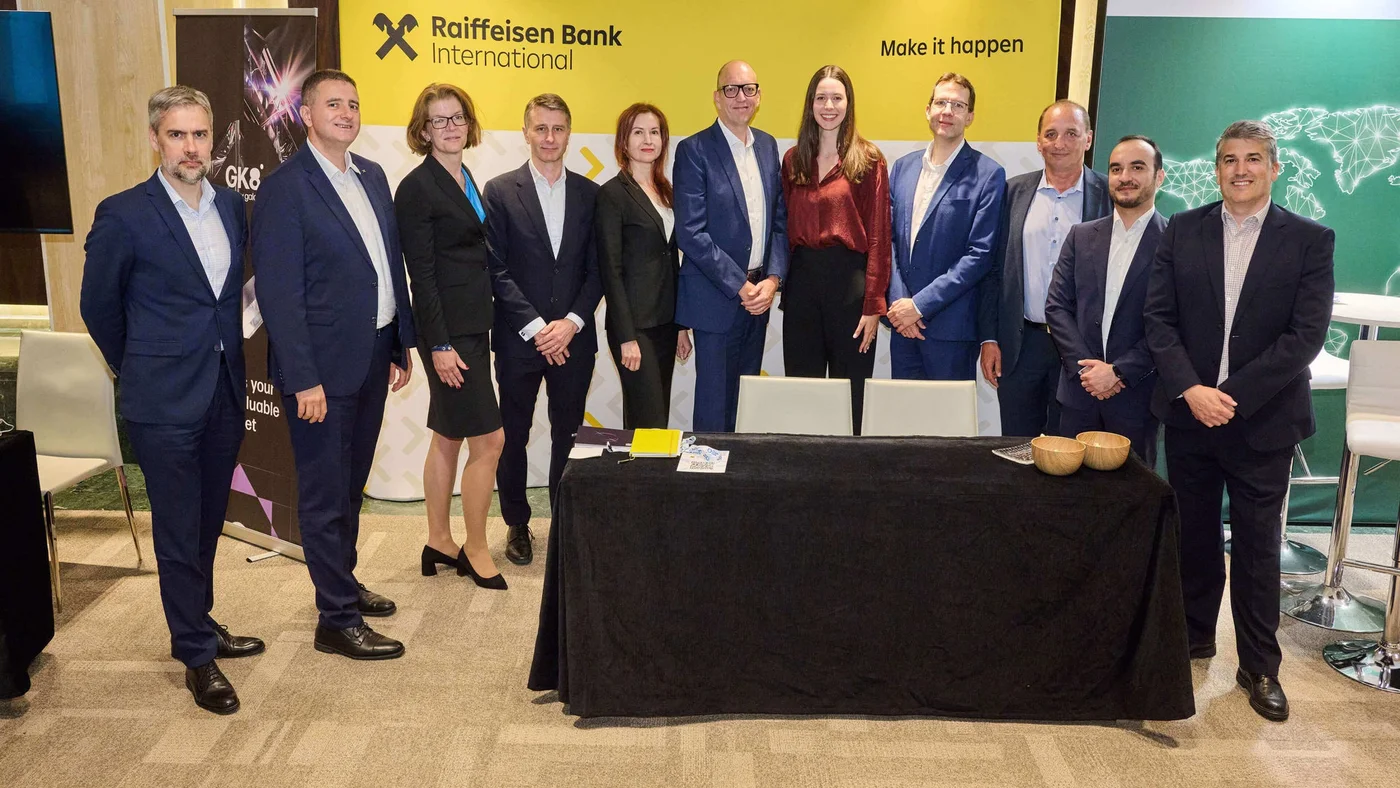 Eine Gruppe von Fachleuten in formeller Kleidung posiert vor einem Banner der Raiffeisen Bank International mit dem Slogan „Make it happen“. Die Szene spielt sich offenbar bei einer Firmenveranstaltung oder Konferenz ab; im Vordergrund steht ein Tisch, im Hintergrund sind weitere Banner zu sehen. Das Bild zeigt eine Mischung aus Männern und Frauen in Businesskleidung und vermittelt eine professionelle, kooperative Atmosphäre.