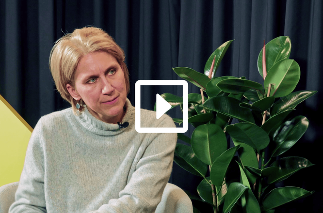 Eine Person sitzt während eines Interviews oder einer Talkshow in einem hellen Pullover, ihr Gesicht ist aus Datenschutzgründen verdeckt. Sie sitzt auf einem hellen Stuhl vor dunklen Vorhängen, wodurch eine studioähnliche Atmosphäre entsteht. Rechts steht eine große, grüne Zimmerpflanze mit glänzenden Blättern. Ein weißes Wiedergabesymbol in der Mitte signalisiert, dass es sich um ein Video-Thumbnail handelt. (wird in einem neuen Tab geöffnet)